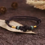 Brown string bracelet with jade bead resting on a dried leaf