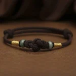 Brown string bracelet with a knot and jade bead resting on a dark dish